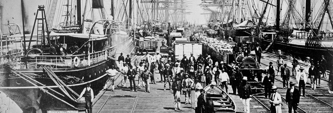 Black and white photo of people on pier with tall ships and pier donkey.